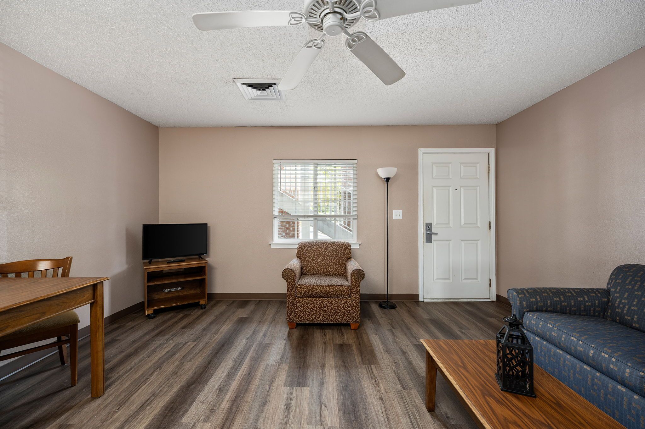 A living room with a wooden table, a patterned armchair, a sofa, a ceiling fan, a TV on a stand, and a window with blinds.