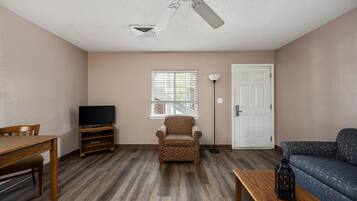 A living room with a wooden table, a patterned armchair, a sofa, a ceiling fan, a TV on a stand, and a window with blinds.