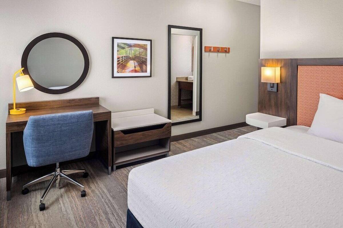 A hotel room with a bed, a desk with a chair, a round mirror, a framed picture, and a nightstand with a lamp.