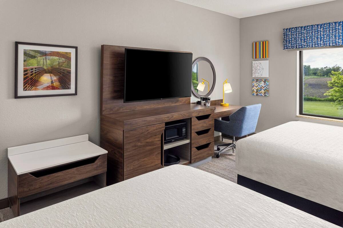 A hotel room with a flat-screen TV, a desk with a chair, a bed, a bedside table, a mirror, and a window with a view of greenery.