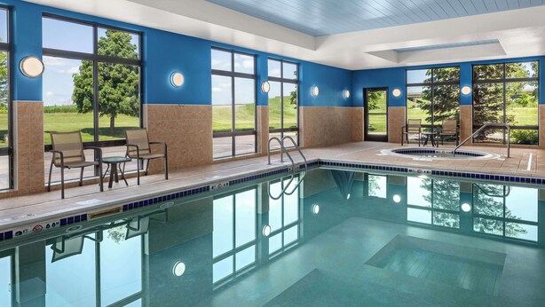 Una piscina interior con paredes de vidrio transparente, vista a un campo cubierto de césped y un área de descanso con sillas y una mesa.