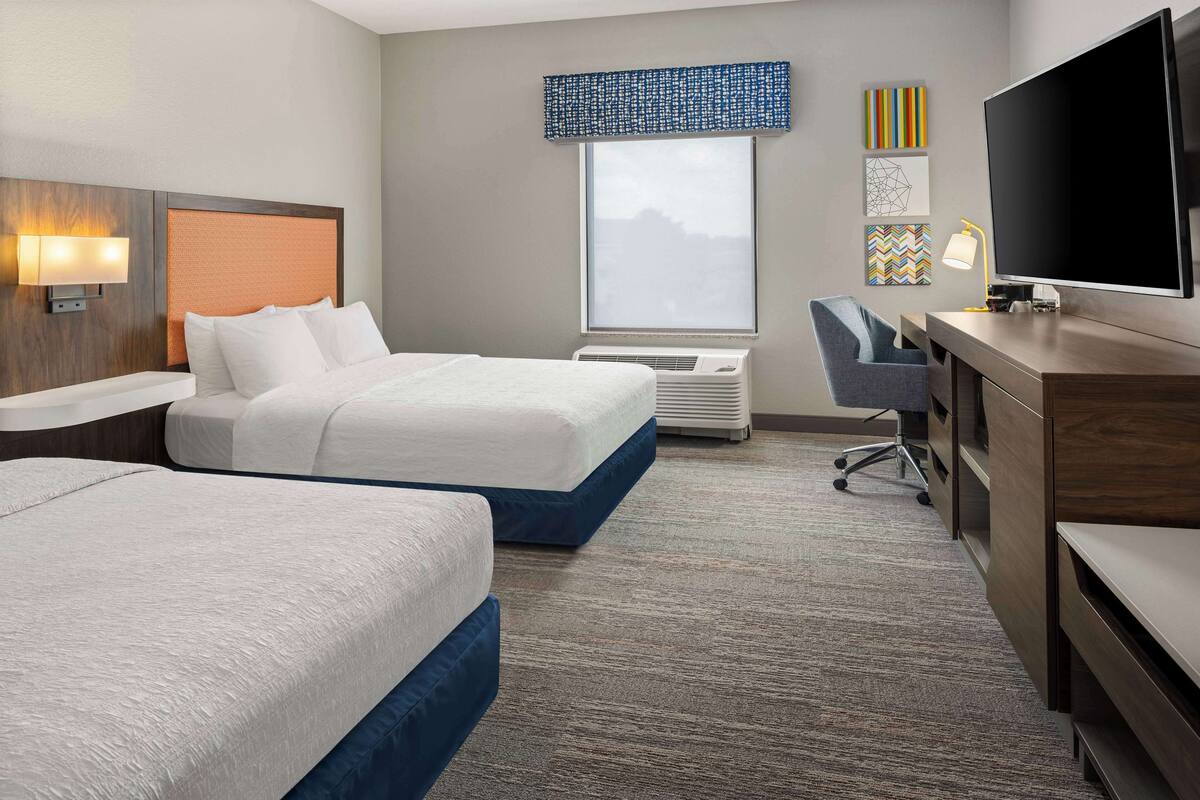 A hotel room with two beds, a desk with a flat-screen TV, a chair, and a window with a view.
