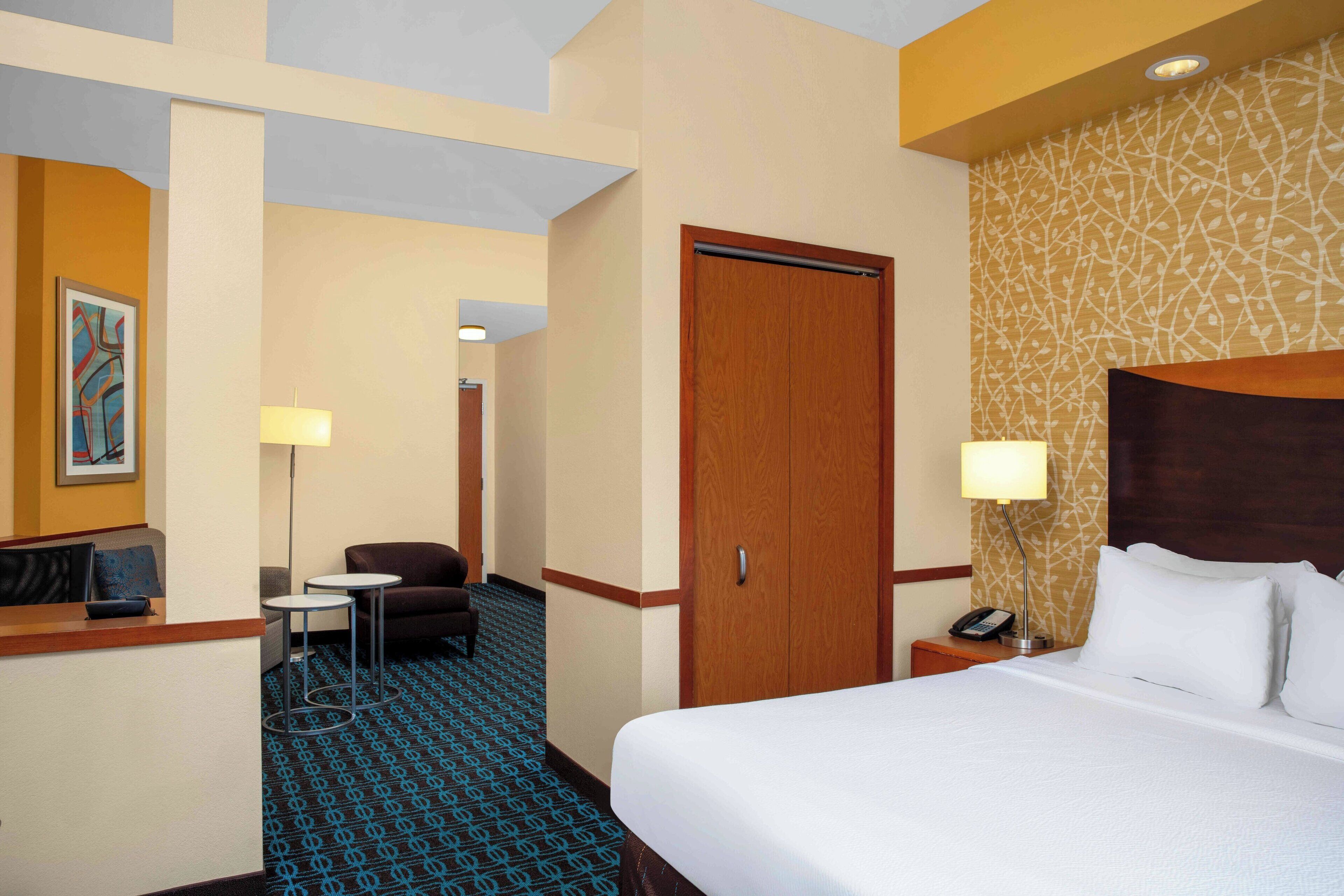 A hotel room with a bed, a desk, a chair, a small table, a lamp, and a phone on the nightstand.