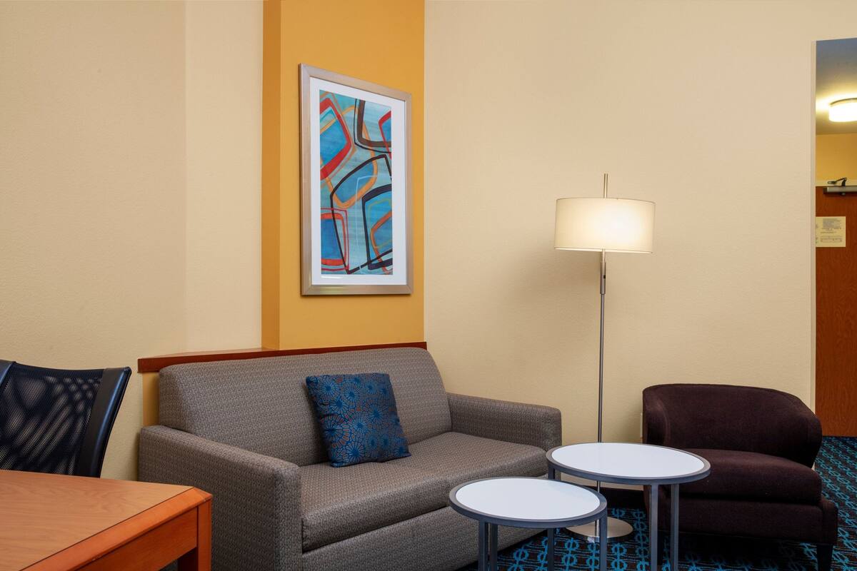 A hotel room with a grey sofa, a blue patterned pillow, two small round tables, a floor lamp, and a framed abstract painting on the wall.