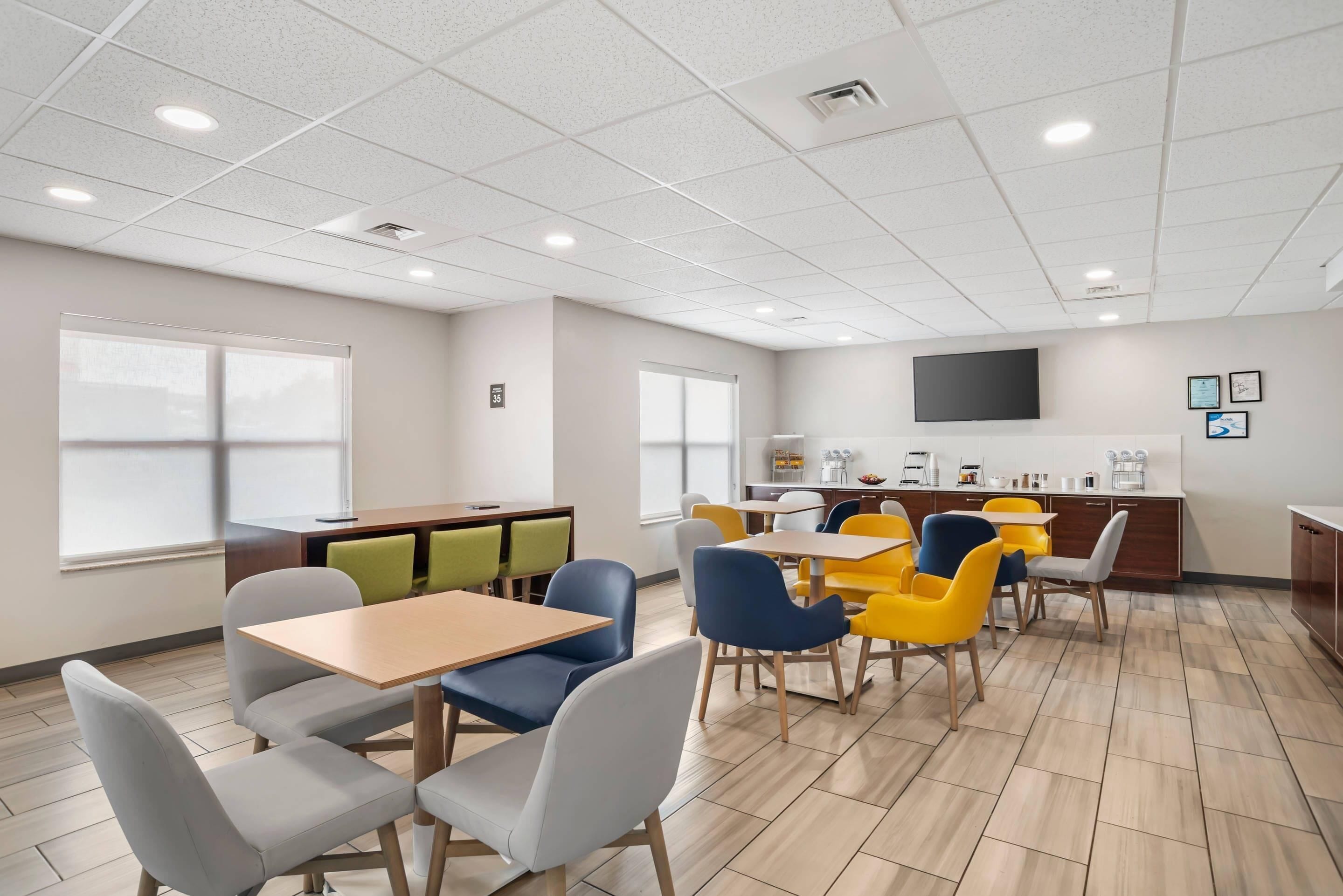 A modern dining area with tables and chairs, a kitchen area with appliances, and a large window providing natural light.