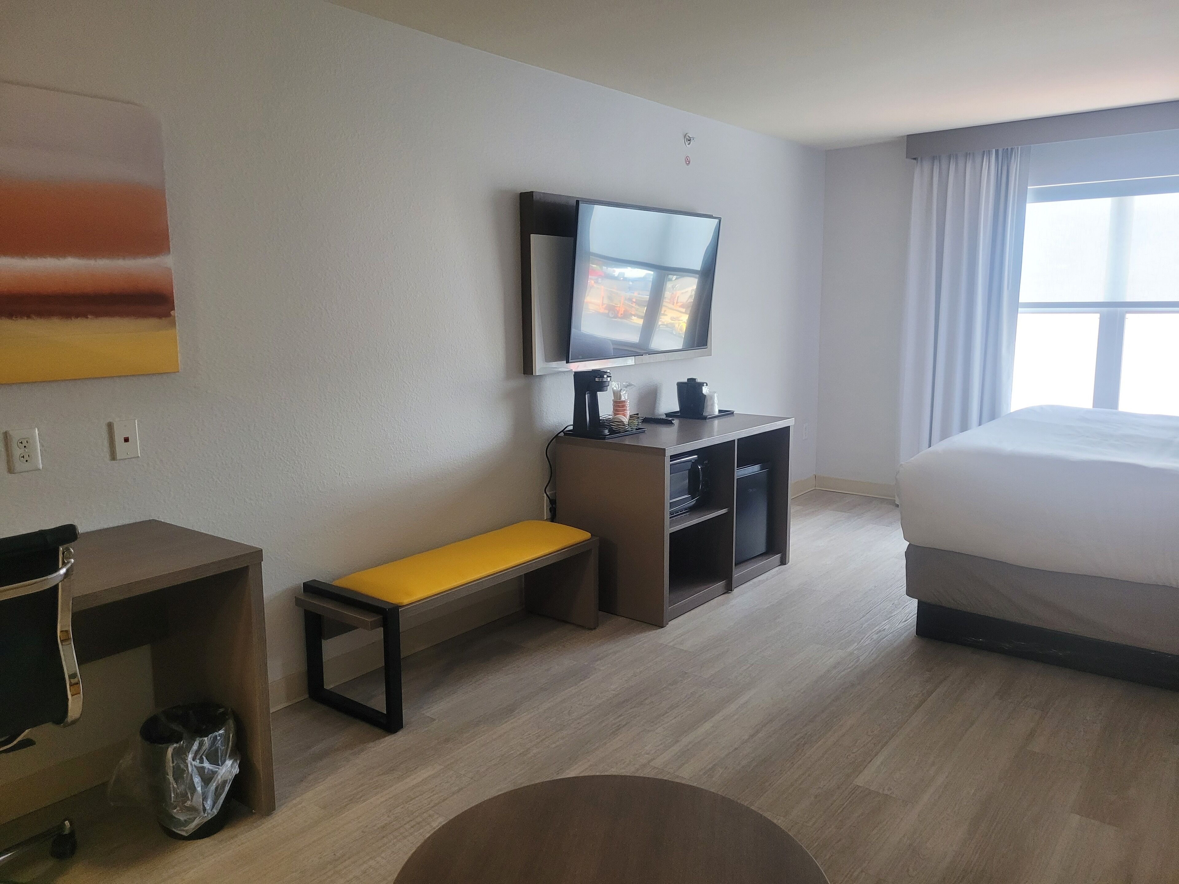 A hotel room with a bed, a desk with a chair, a bench, a TV mounted on the wall, and a window with curtains.