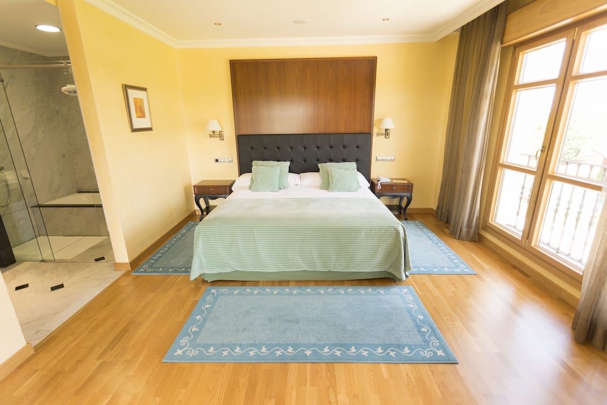 Ein Hotelzimmer mit einem großen Bett, zwei Nachttischen, einem Holzkopfteil, einer Duschkabine und einem großen Fenster mit Vorhängen.