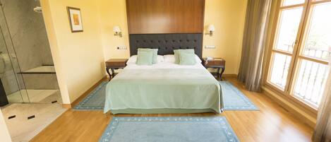 A hotel room with a large bed, two bedside tables, a wooden headboard, a shower area, and a large window with curtains.