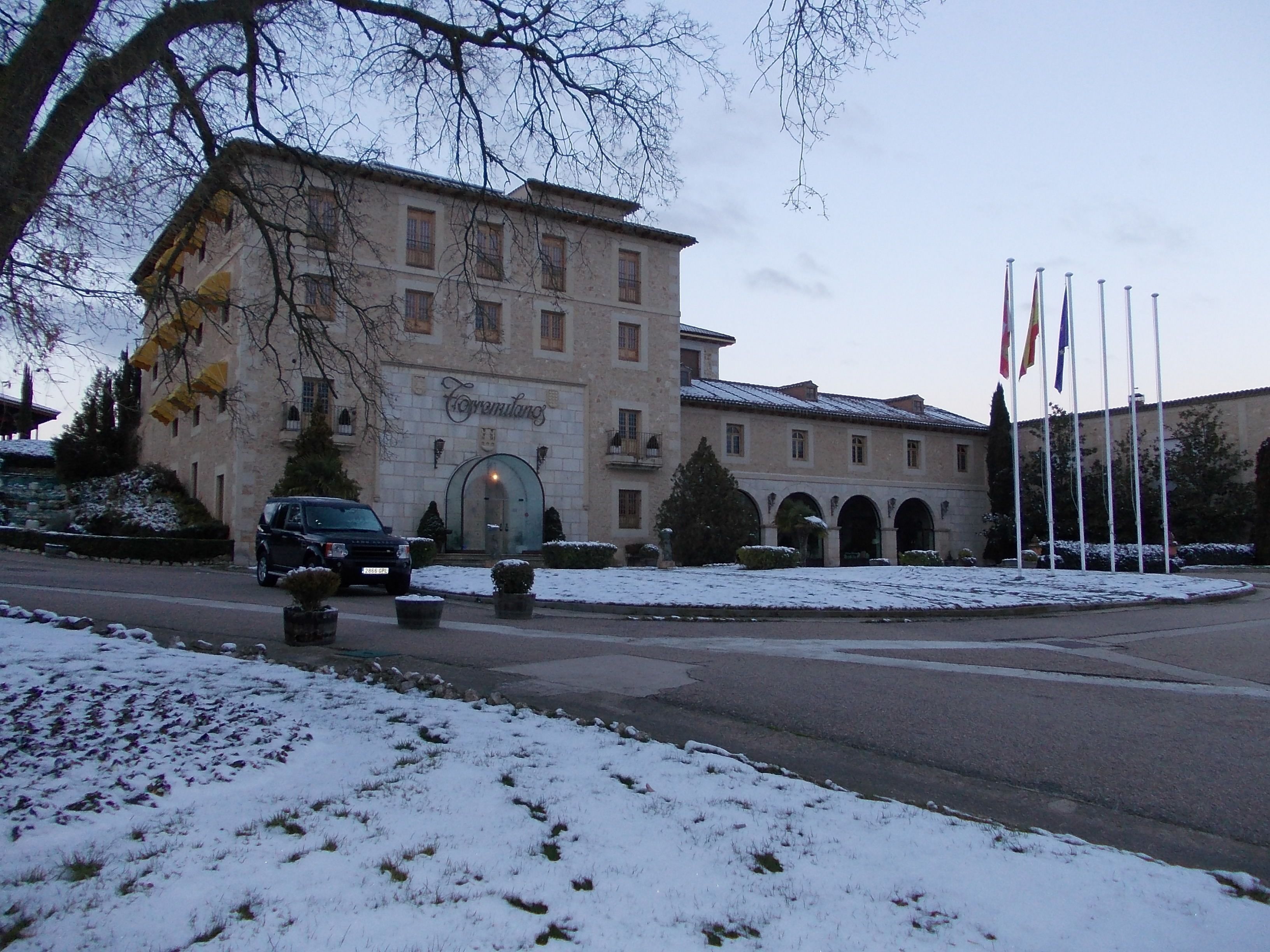 A historic building with a stone facade, a central entrance, and multiple windows. There are flags in front of the building, and snow on the ground.