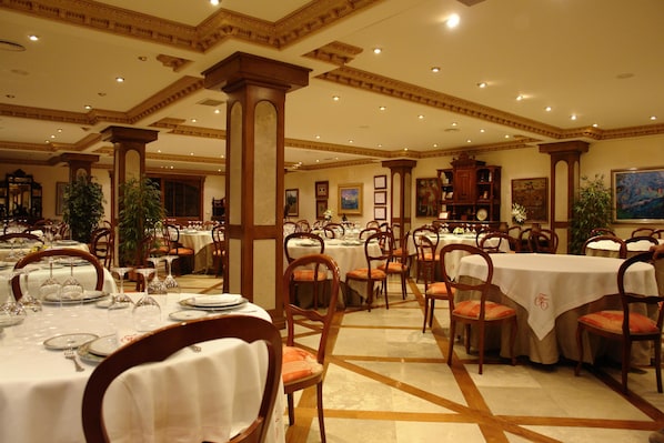 A spacious dining area with multiple tables and chairs, elegant ceiling design, and framed artwork on the walls.