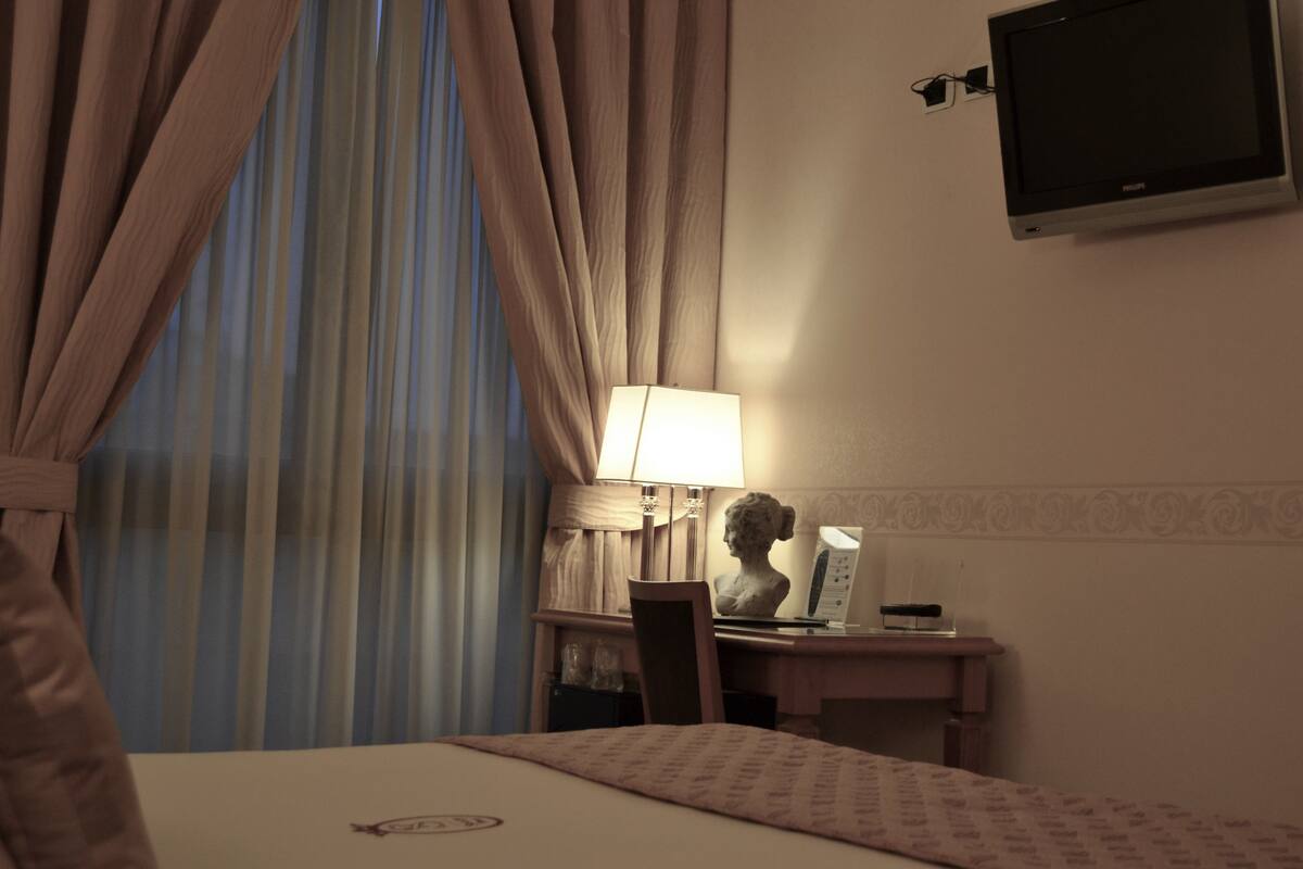 A hotel room with a bed, a desk, a lamp, a bust sculpture, and a television on the wall.