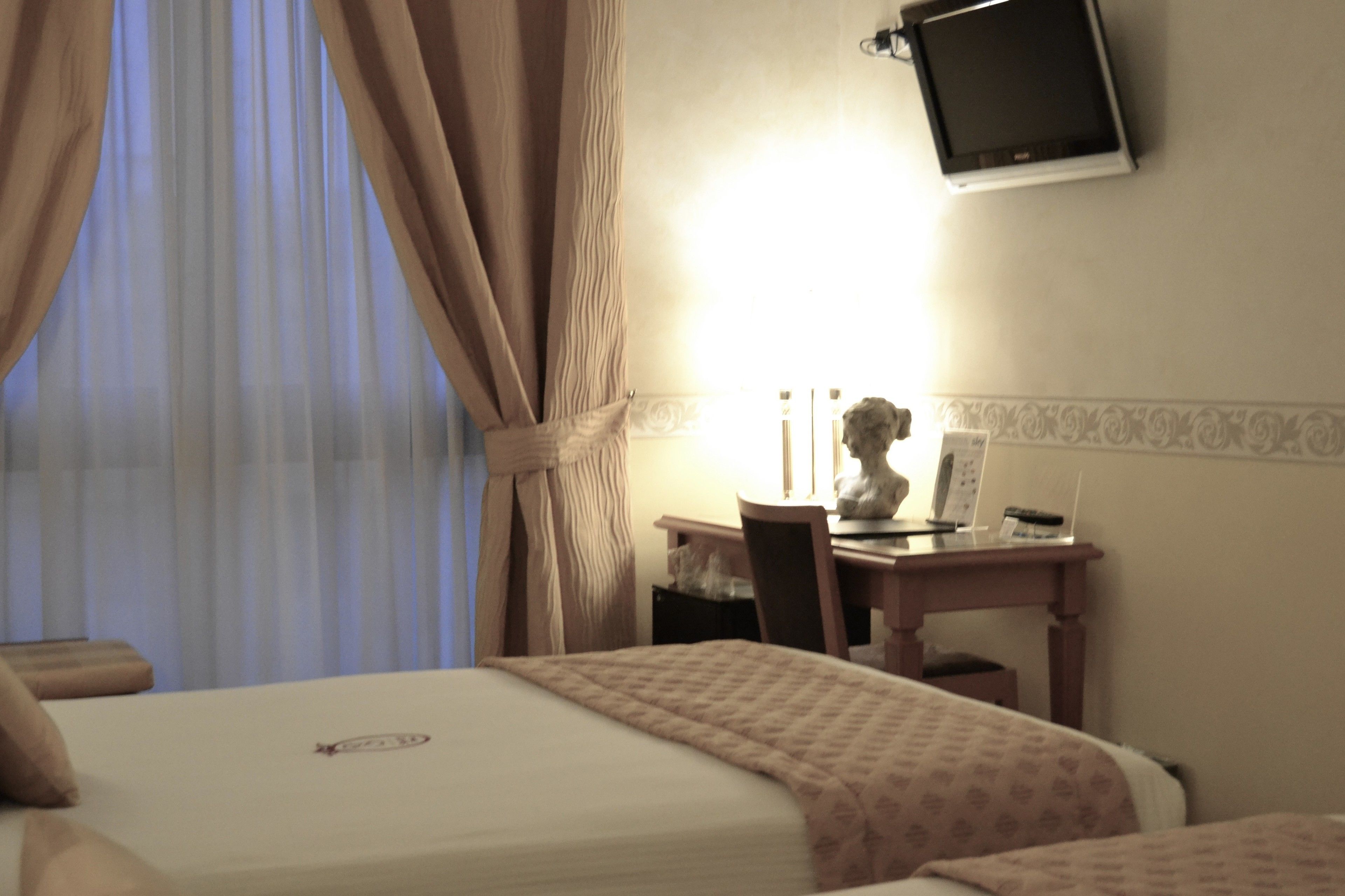 A hotel room with a bed, a desk, a chair, a television mounted on the wall, and a window with curtains.