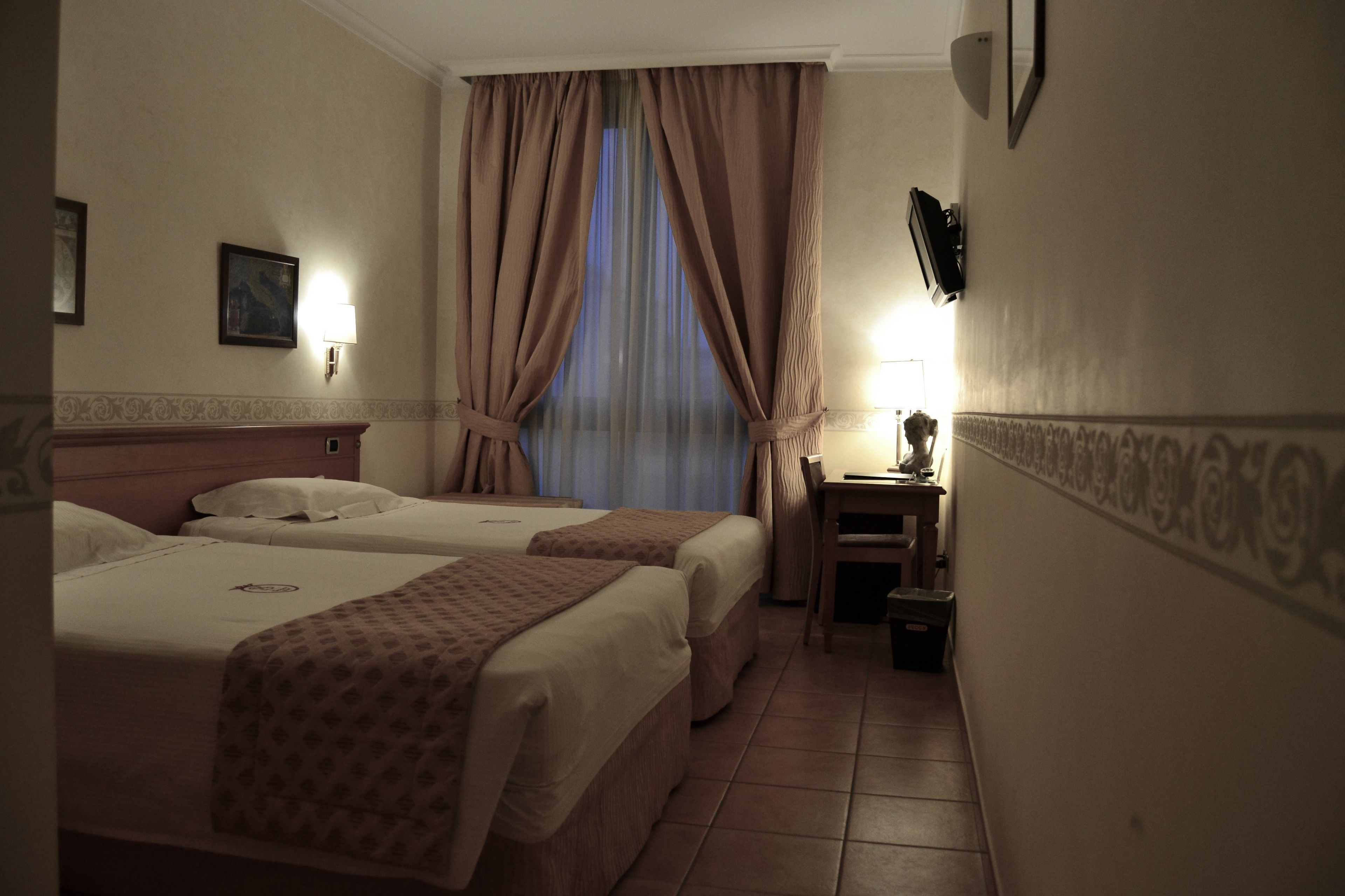 Quarto de hotel com duas camas, uma mesinha com uma luminária, uma cadeira e uma janela com cortinas.
