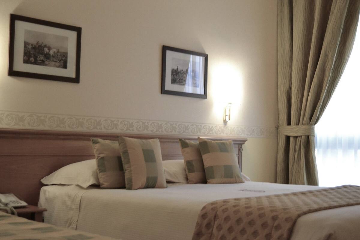 A hotel room with a bed, two framed pictures on the wall, a nightstand, and a window with curtains.