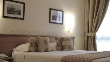 A hotel room with a bed, two framed pictures on the wall, a nightstand, and a window with curtains.