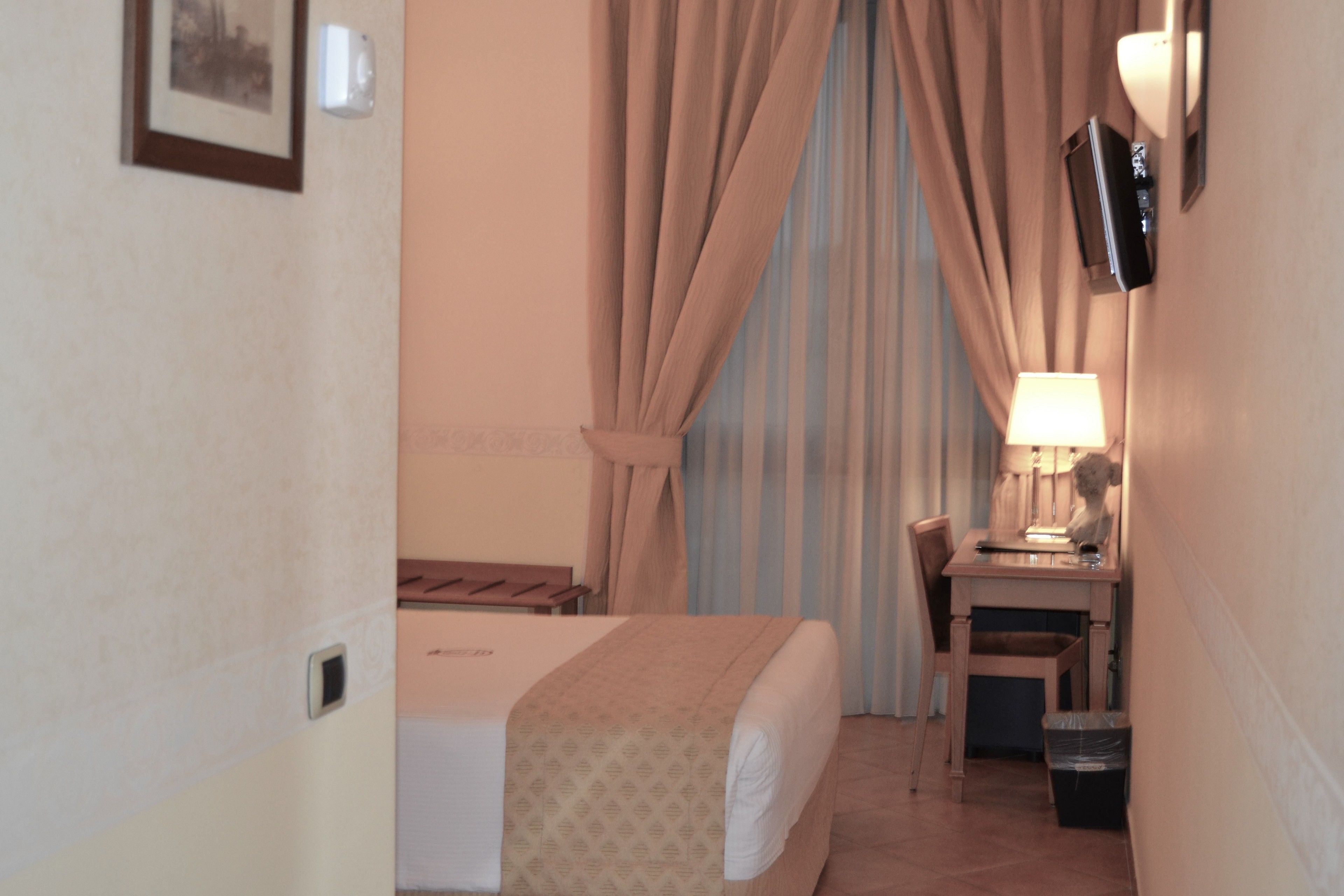 A hotel room with a bed, a desk, a chair, a lamp, and a television on the wall.