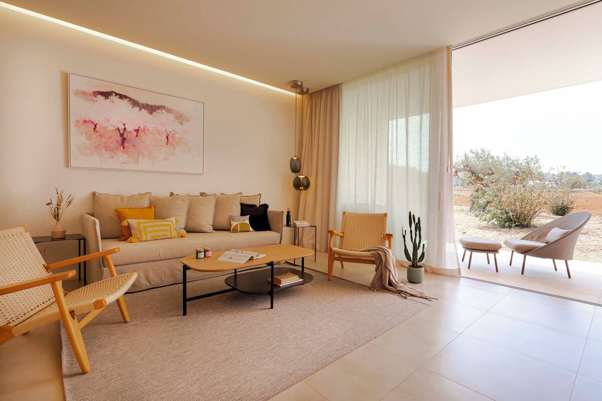 Een moderne woonkamer met een bank, salontafel en twee stoelen. Er is een groot raam met gordijnen, een cactuss en een schilderij aan de muur.
