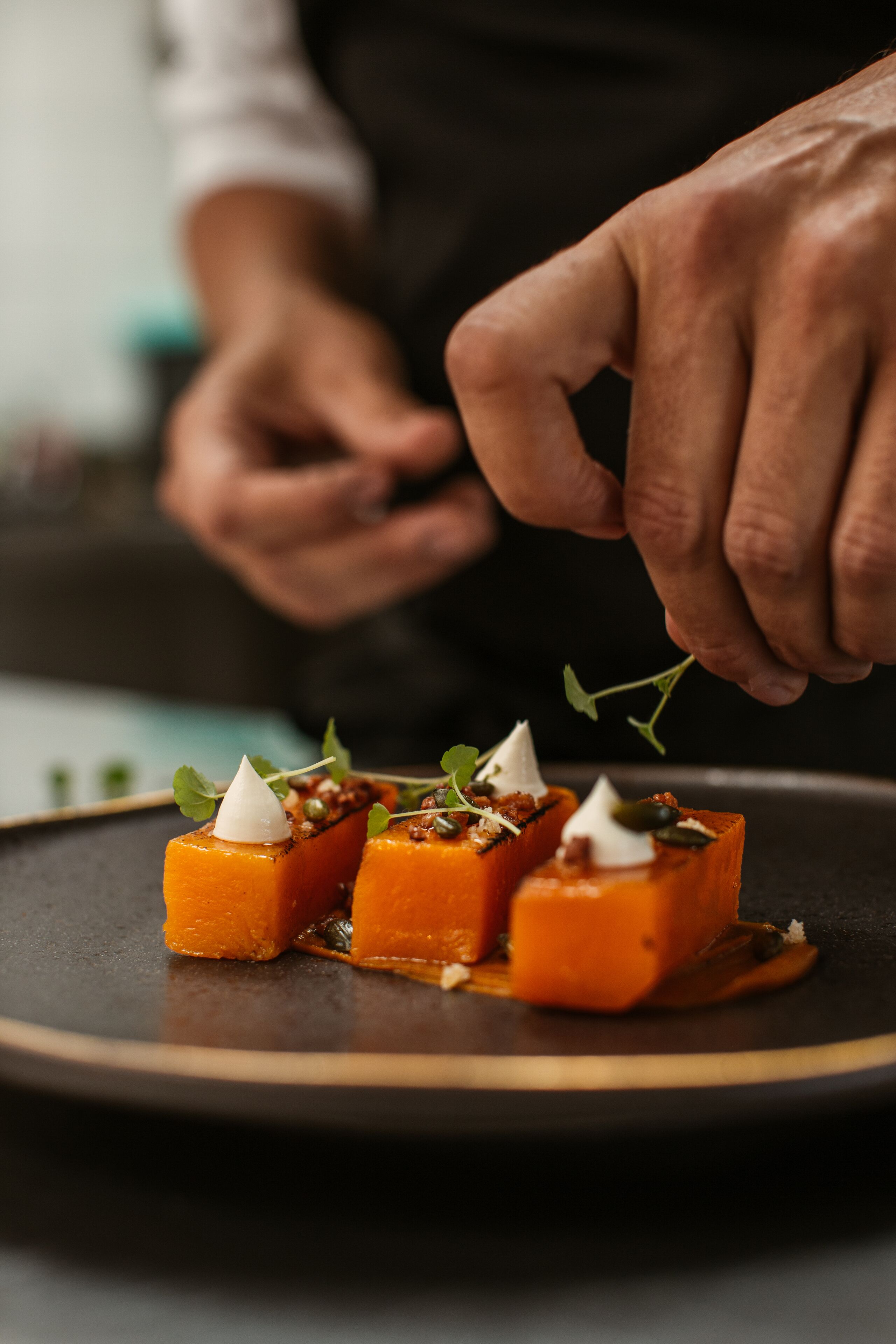 Les mains d’un chef décorant un plat avec une sauce blanche et des herbes.