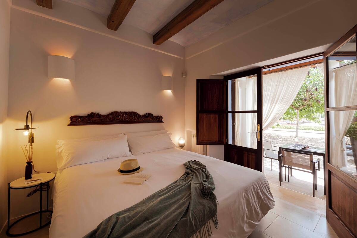Een slaapkamer met een groot bed, bedlampjes, een klein tafeltje met een lamp, en uitzicht op een terras met een tafel en stoelen.