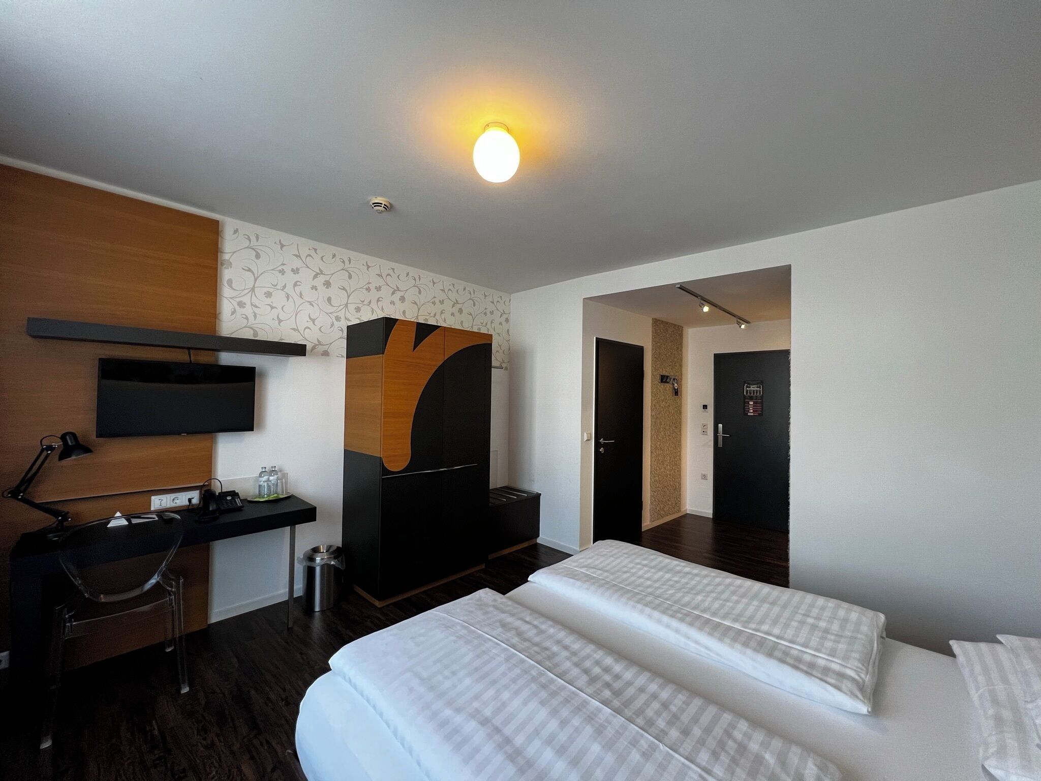 A hotel room with a bed, a desk with a computer, a TV, a wardrobe, and a door leading to another room.