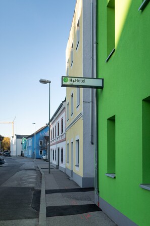 A street view with a green building labeled "H+Hotel" and adjacent buildings in blue and yellow.