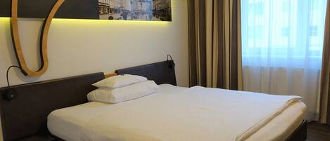 Habitación de hotel con una cama grande, cabecera con lámpara, una obra de arte colgada en la pared y una ventana con cortinas.