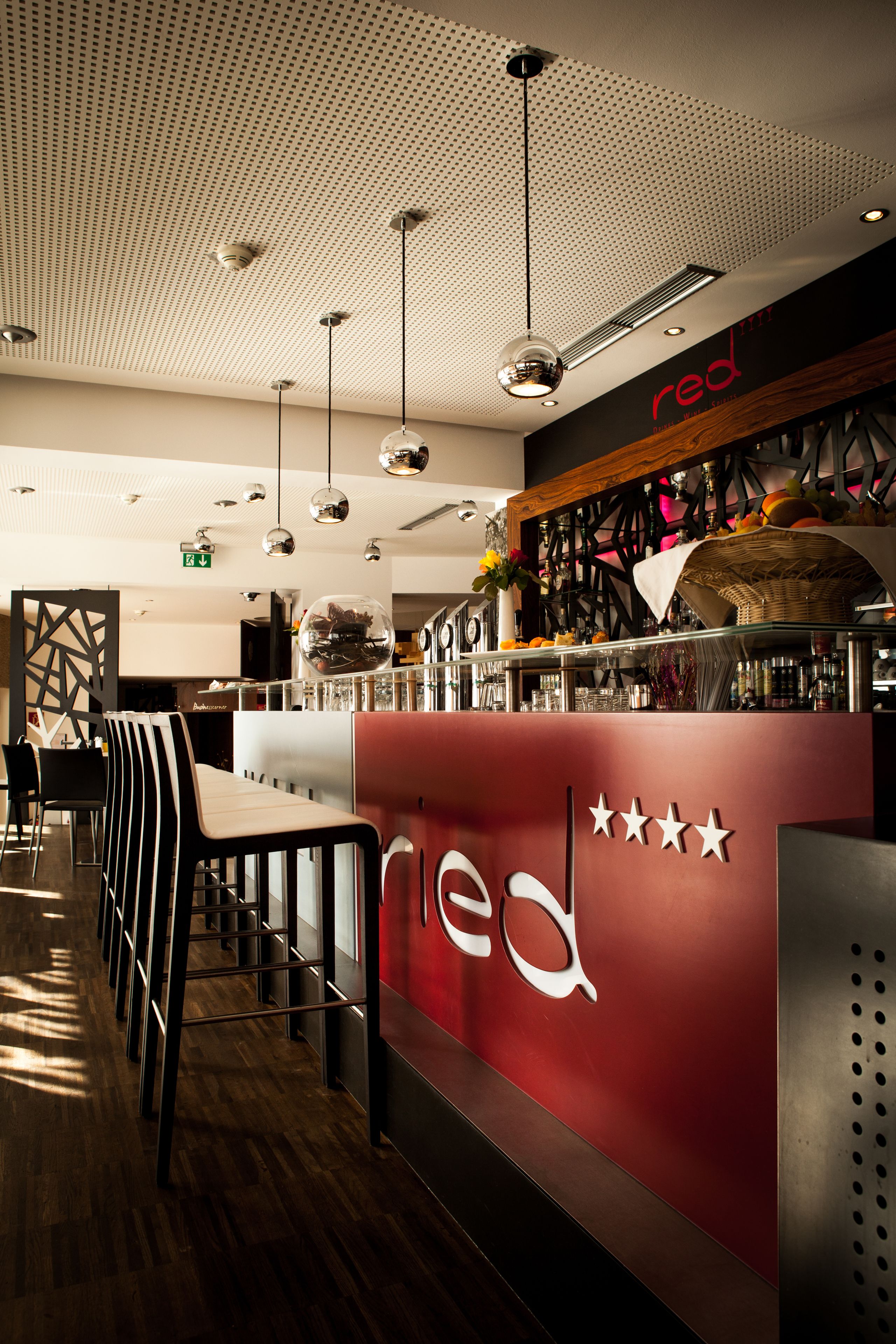 A modern bar area with a red and black color scheme, featuring a bar counter, multiple hanging pendant lights, and a ceiling with a perforated design.
