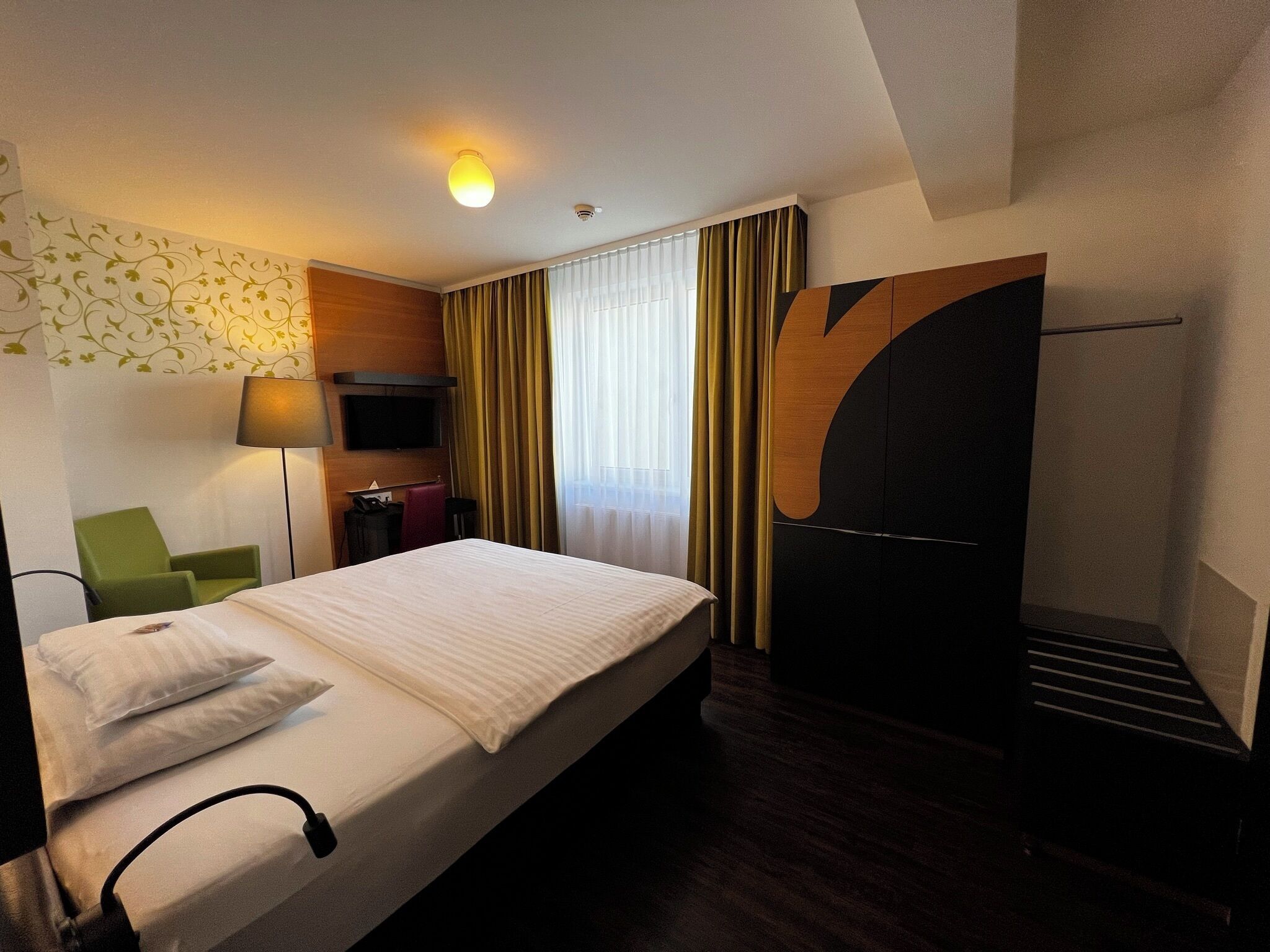 A hotel room with a bed, a TV, a desk, a chair, and a large window with curtains.