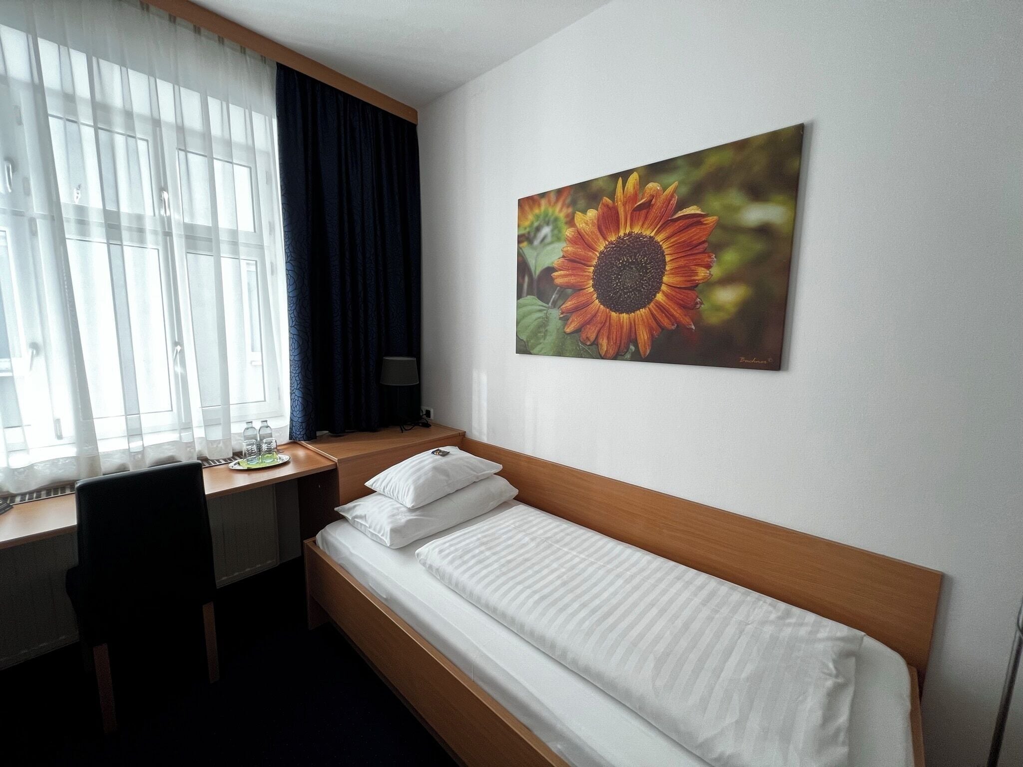 A hotel room with a bed, a desk, a chair, a window with curtains, and a painting of a sunflower.