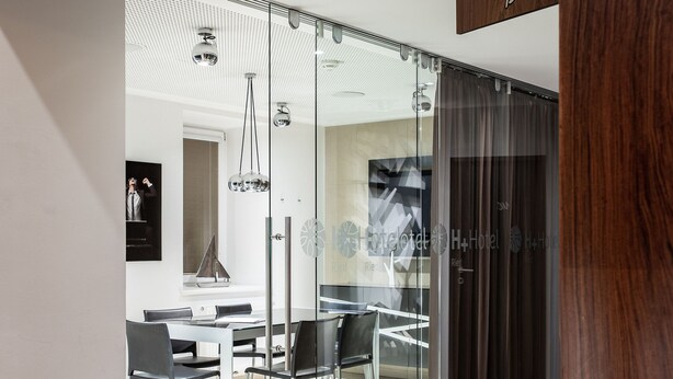 A modern hotel room with a glass partition, a dining area with a white table and black chairs, and a wooden accent wall with the word "Business". - H+ Hotel Ried (Ried im Innkreis)