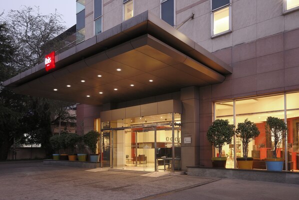 Fachada de hotel de vidrio, zona de acceso techada y plantas en macetas.