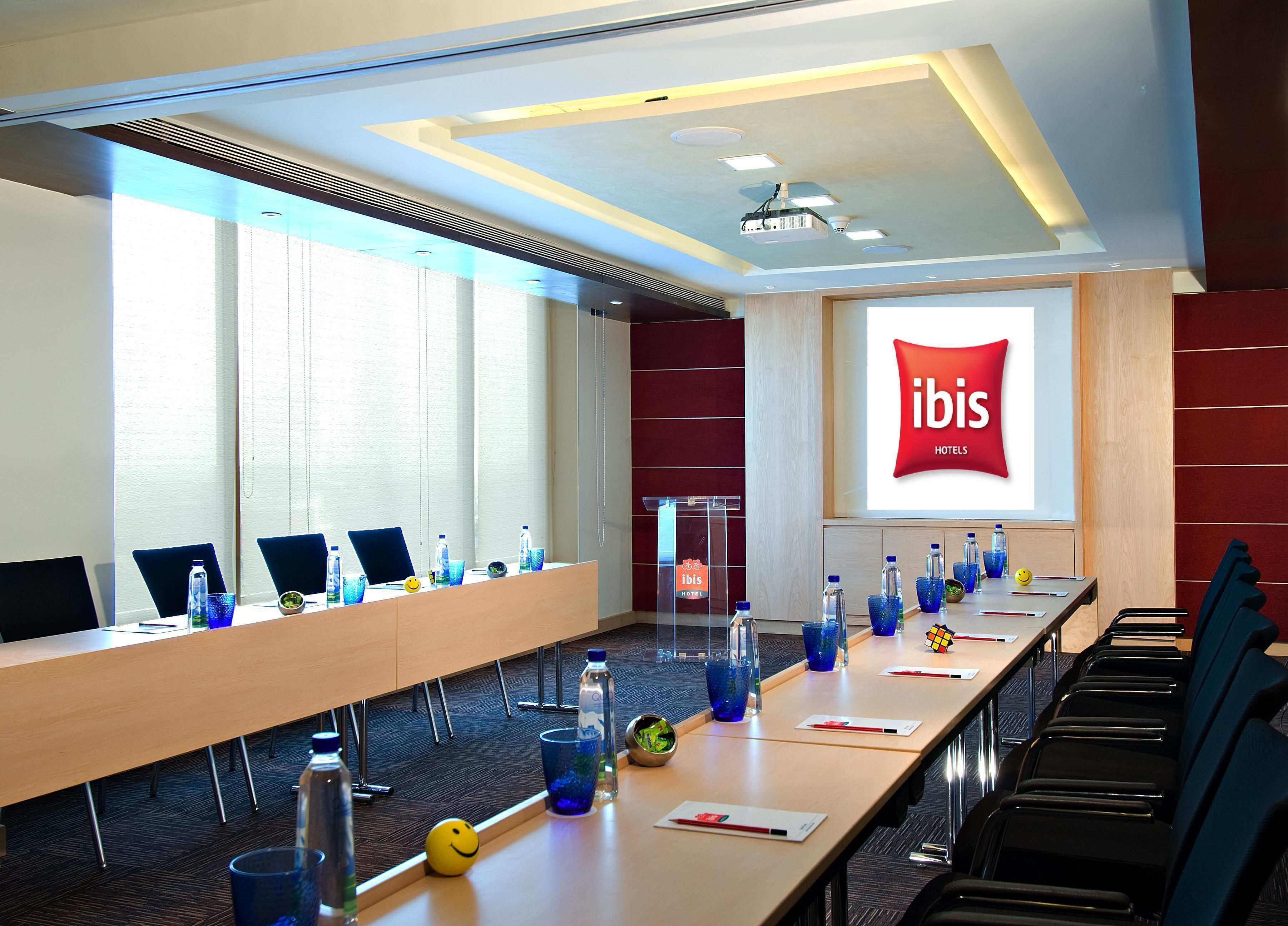 A conference room with a long table, chairs, and a projector screen displaying the ibis hotel logo.