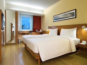 Ett hotellrum med två sängar, ett skrivbord, en stol, en spegel, ett fönster och en inramad tavla på väggen.