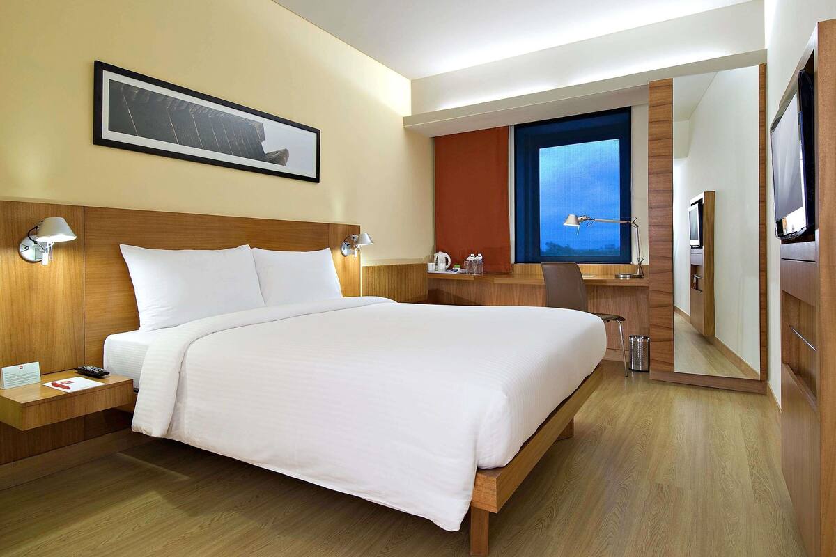A hotel room with a bed, bedside tables, a desk, a chair, a TV, and a window.