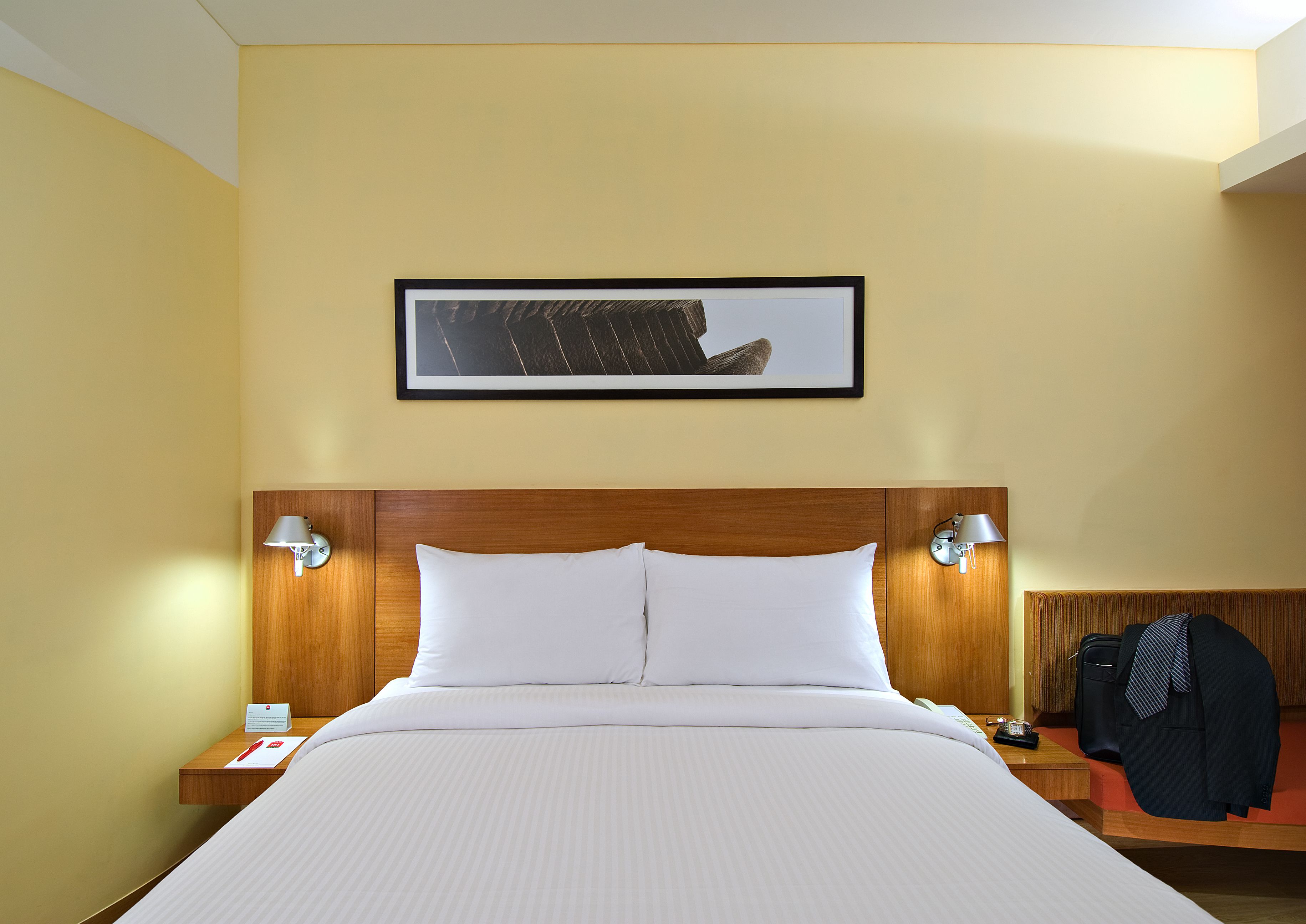 Una habitació d'hotel amb un llit gran, dues taules de nit, un quadre al mur i un penjador amb una jaqueta.