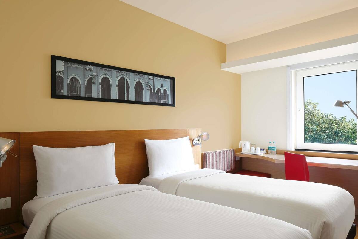 A hotel room with two beds, a window with a view, a desk with a lamp, and a framed picture on the wall.