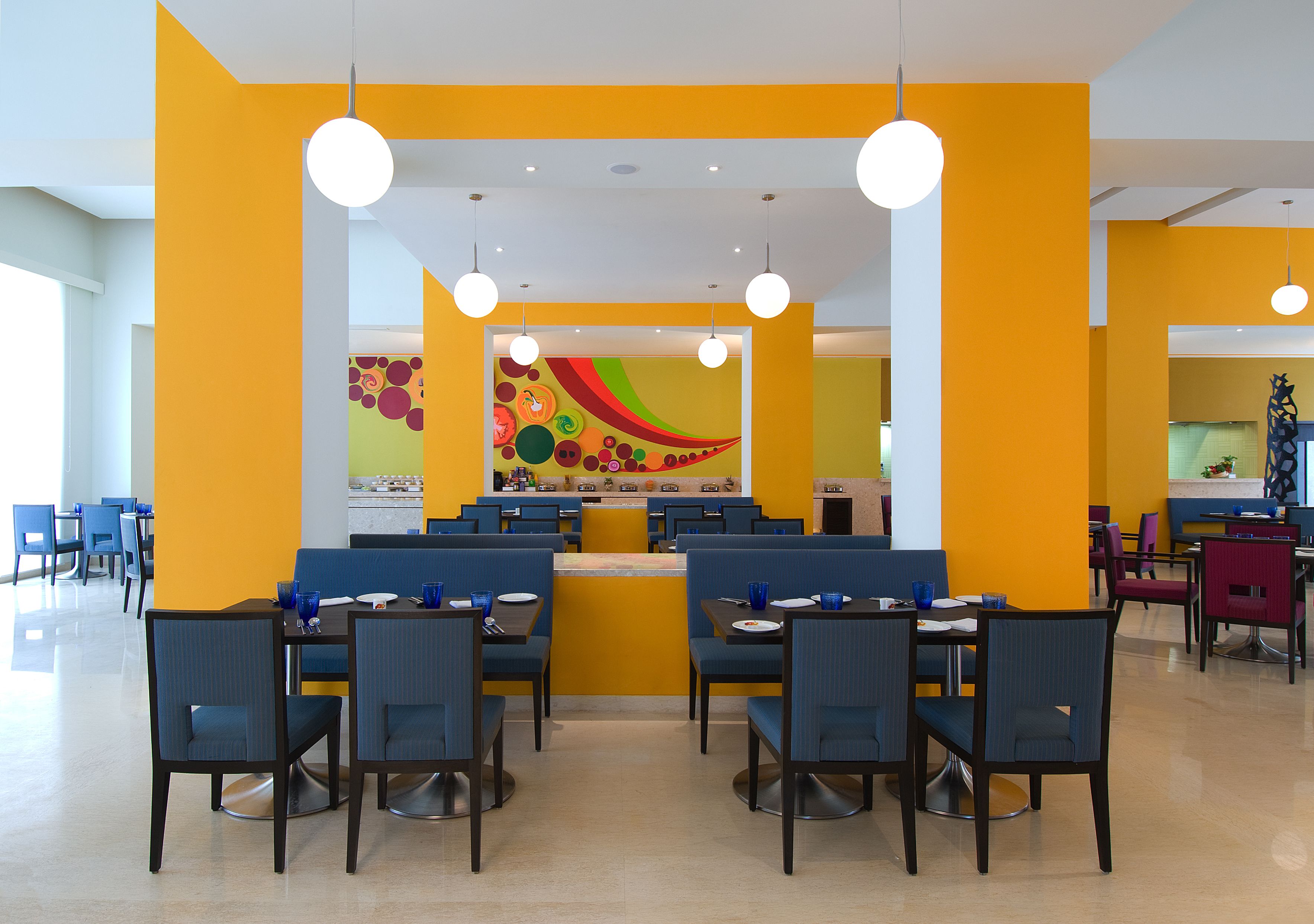 En moderne restaurant med gule vægge, blå og røde stole og et farverigt mural på bagvæggen.