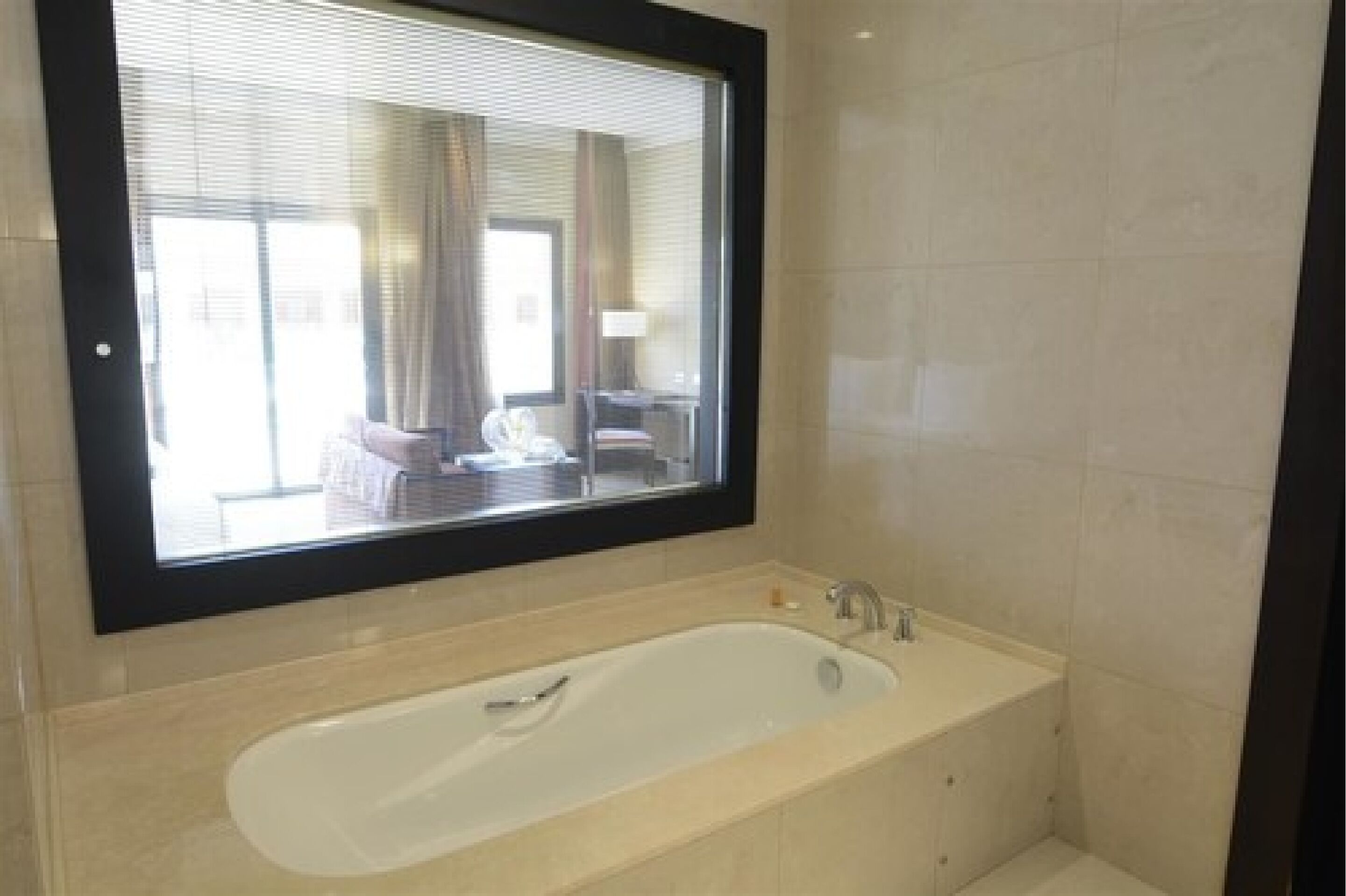 A modern bathroom with a large bathtub, a mirror above the tub, and a view of a bedroom through the window.