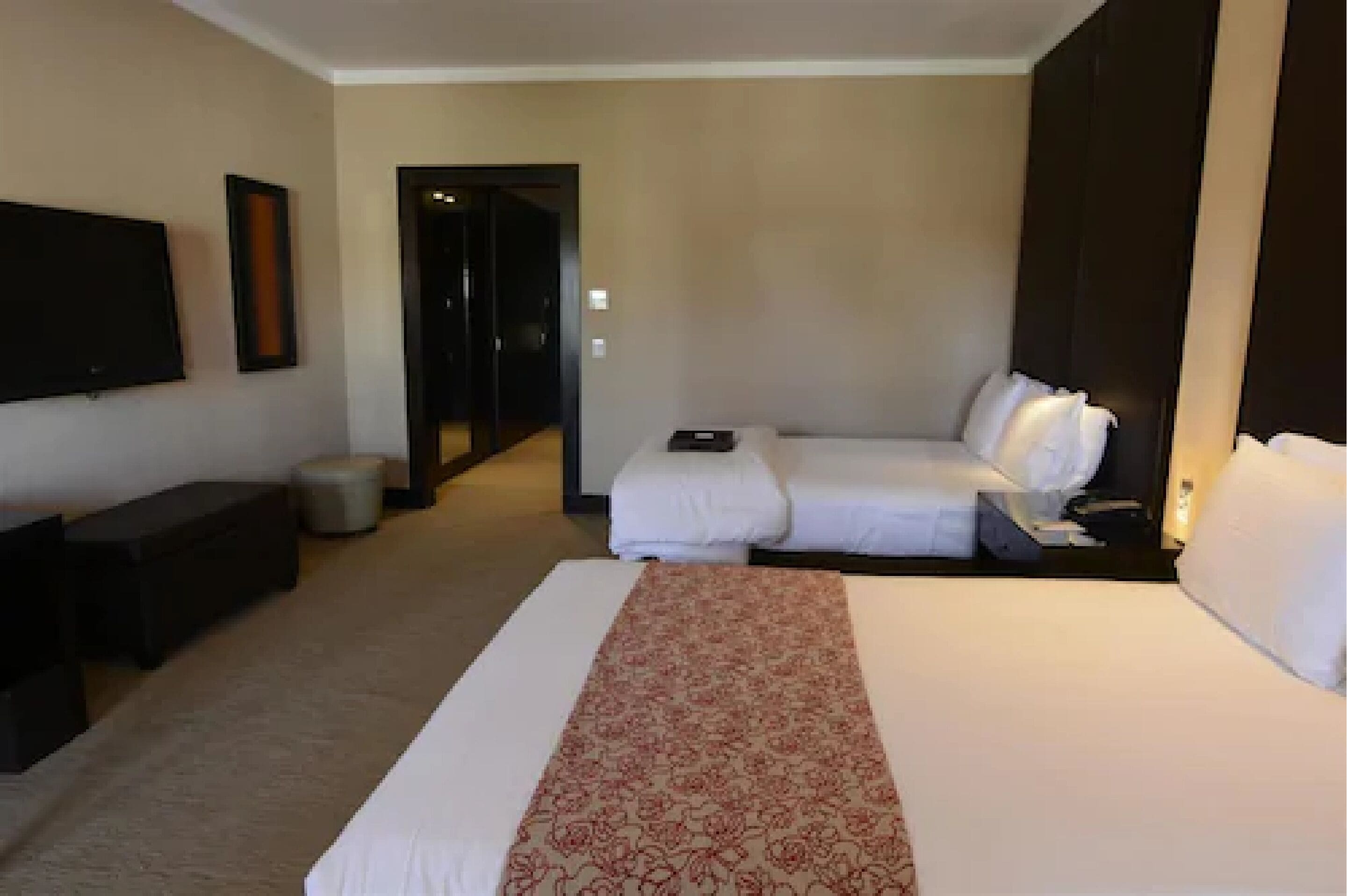 Habitación de hotel con una cama grande, una cama más pequeña, un televisor, un escritorio y una puerta que da a un pasillo.