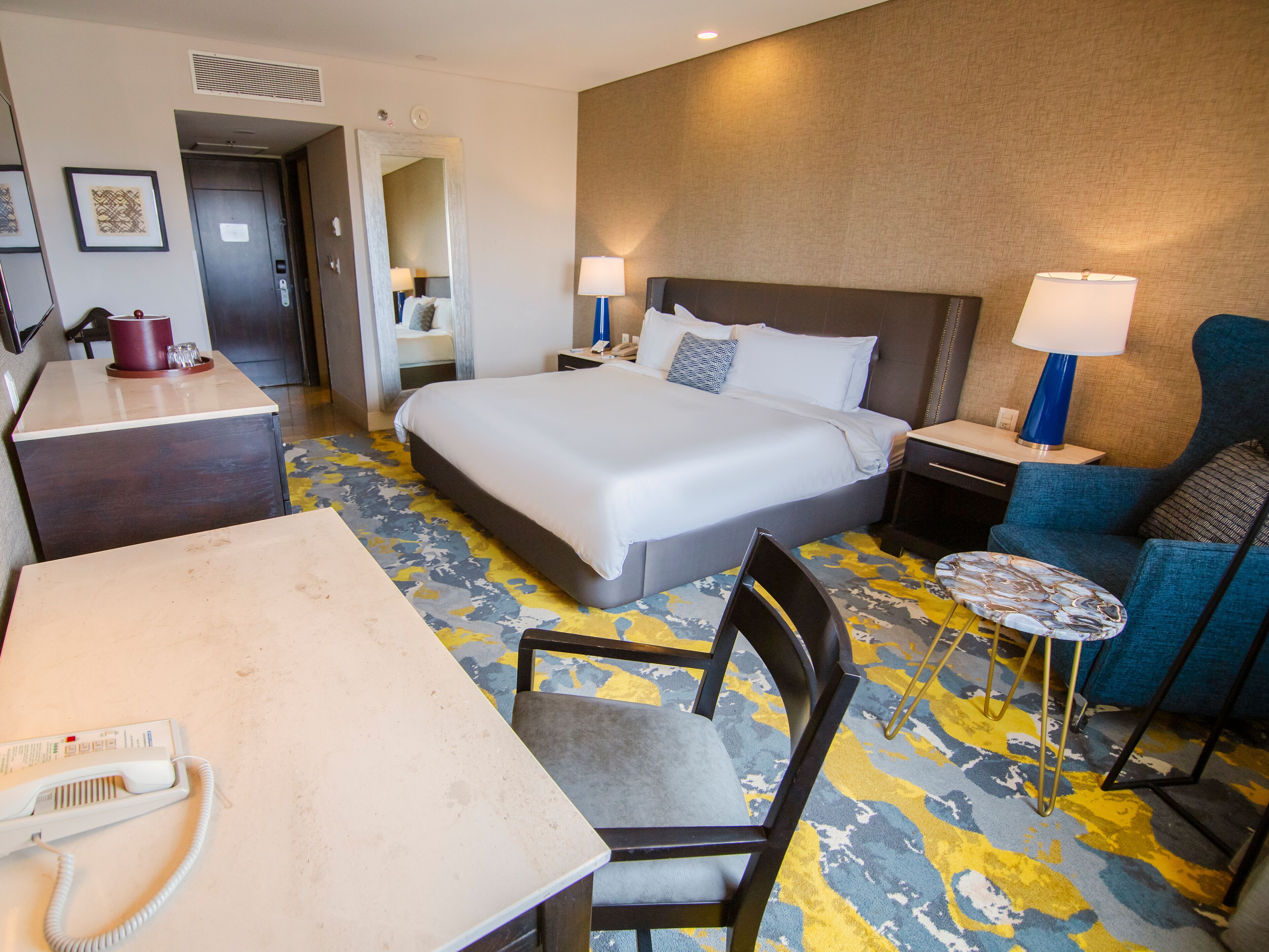 Quarto de hotel com uma cama grande, uma escrivaninha com telefone, uma cadeira, uma mesinha de cabeceira com abajur e uma poltrona azul.