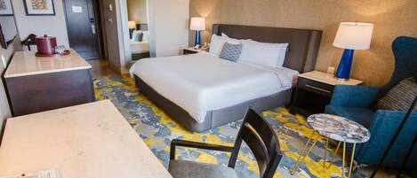 Quarto de hotel com uma cama grande, uma escrivaninha com telefone, uma cadeira, uma mesinha de cabeceira com abajur e uma poltrona azul.