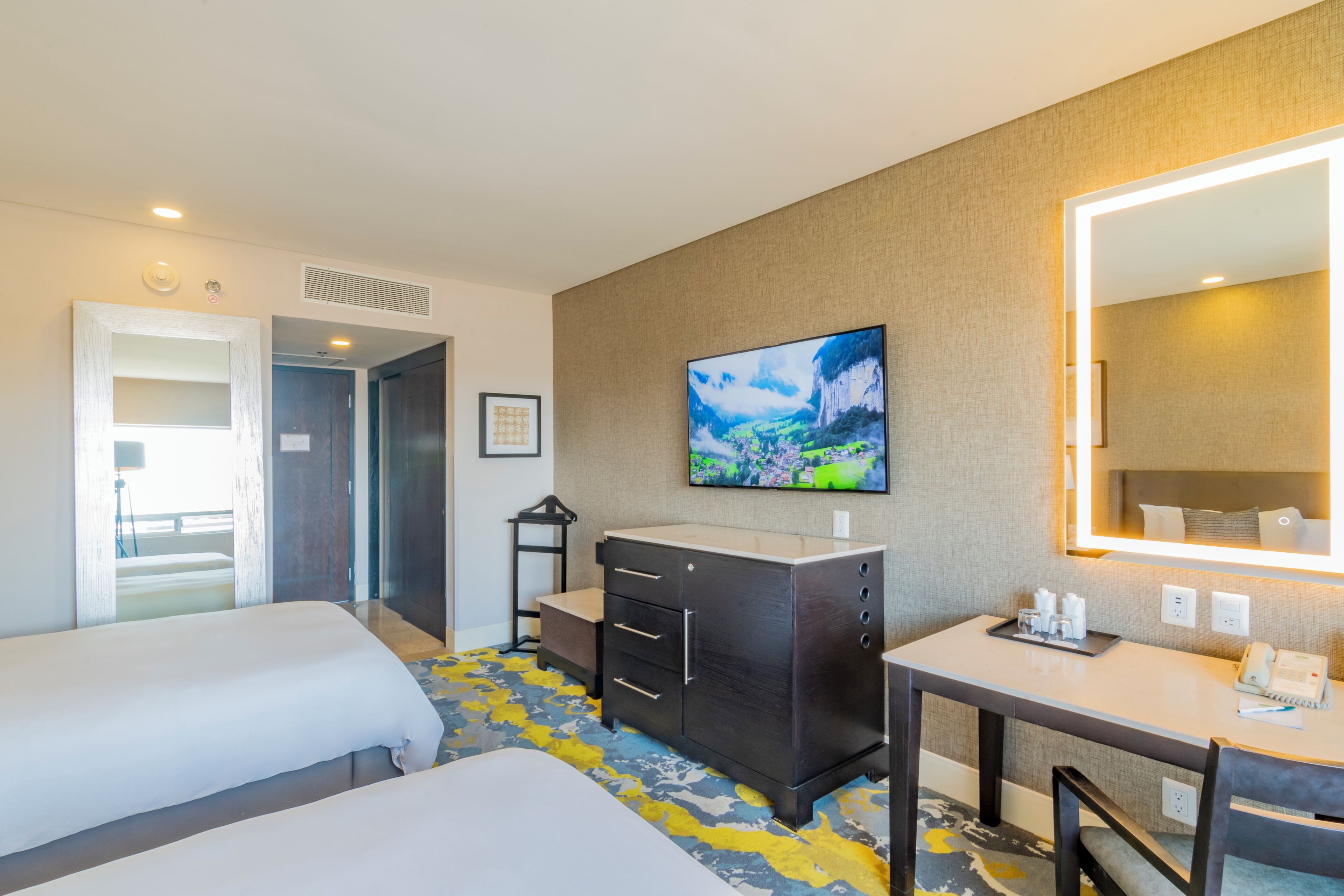 A hotel room with two beds, a desk, a dresser, a TV, and a large mirror.