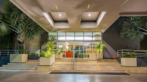 A modern hotel entrance with a glass facade, a staircase leading to the lobby, and potted plants on either side.