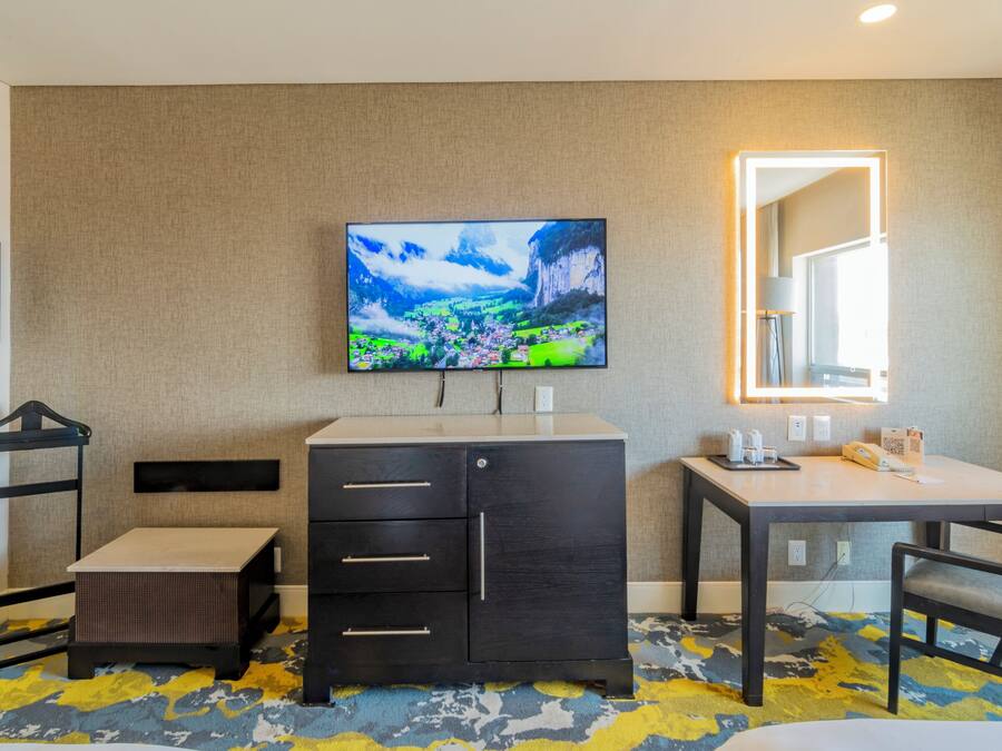 Habitación de hotel con televisor de pantalla plana en la pared, un escritorio con silla, un armario pequeño y una cama.