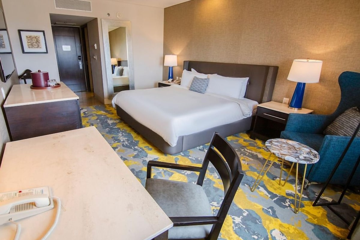 A hotel room with a bed, bedside tables, a desk with a telephone, a chair, a small round table, and a blue armchair.