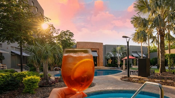 A hand holding a glass with a slice of orange, a swimming pool, palm trees, and a building in the background.