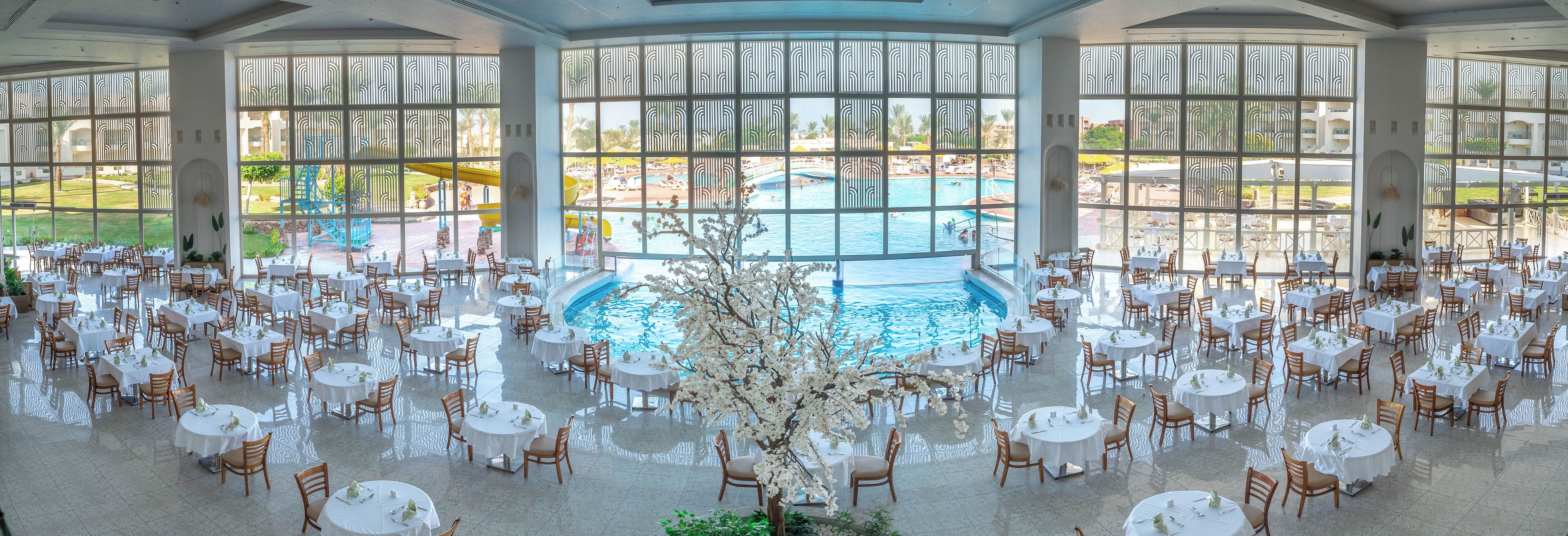 Un vaste espace de restauration intérieur doté de tables et de chaises rondes, d’une piscine centrale et de grandes fenêtres offrant une vue sur une piscine extérieure et un jardin.