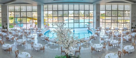 A spacious indoor dining area with round tables and chairs, a central pool, and large windows offering views of an outdoor pool and garden.