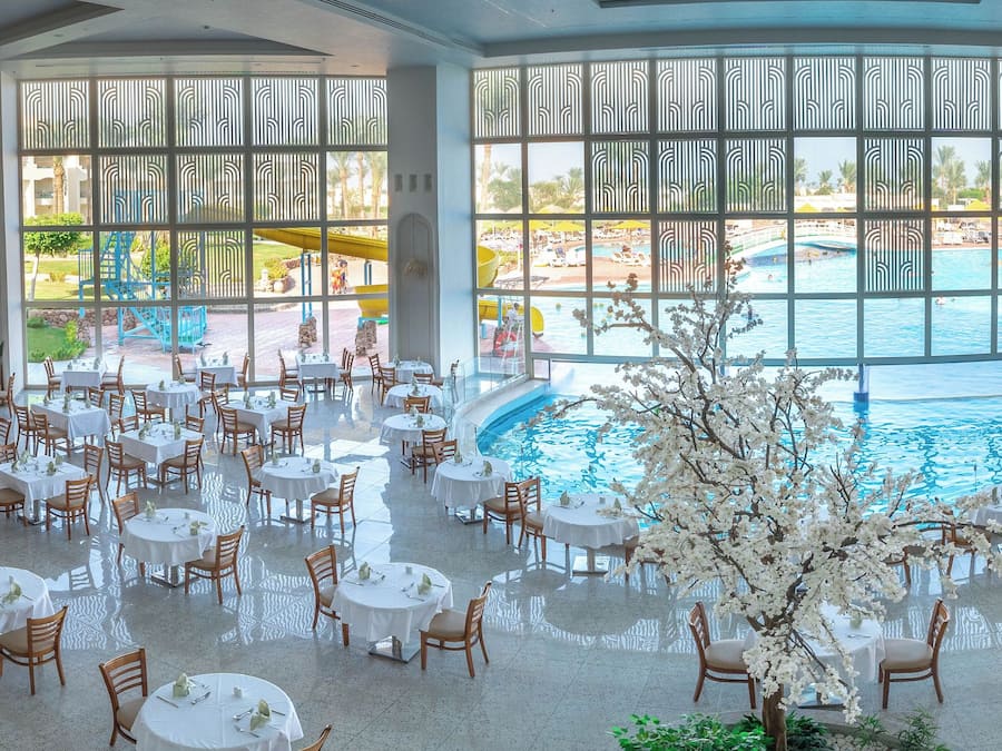 Ampla área de refeições interna, com mesas e cadeiras redondas, piscina central e grandes janelas com vista para uma piscina e jardim externos.