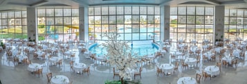 A spacious indoor dining area with round tables and chairs, a central pool, and large windows offering views of an outdoor pool and garden.